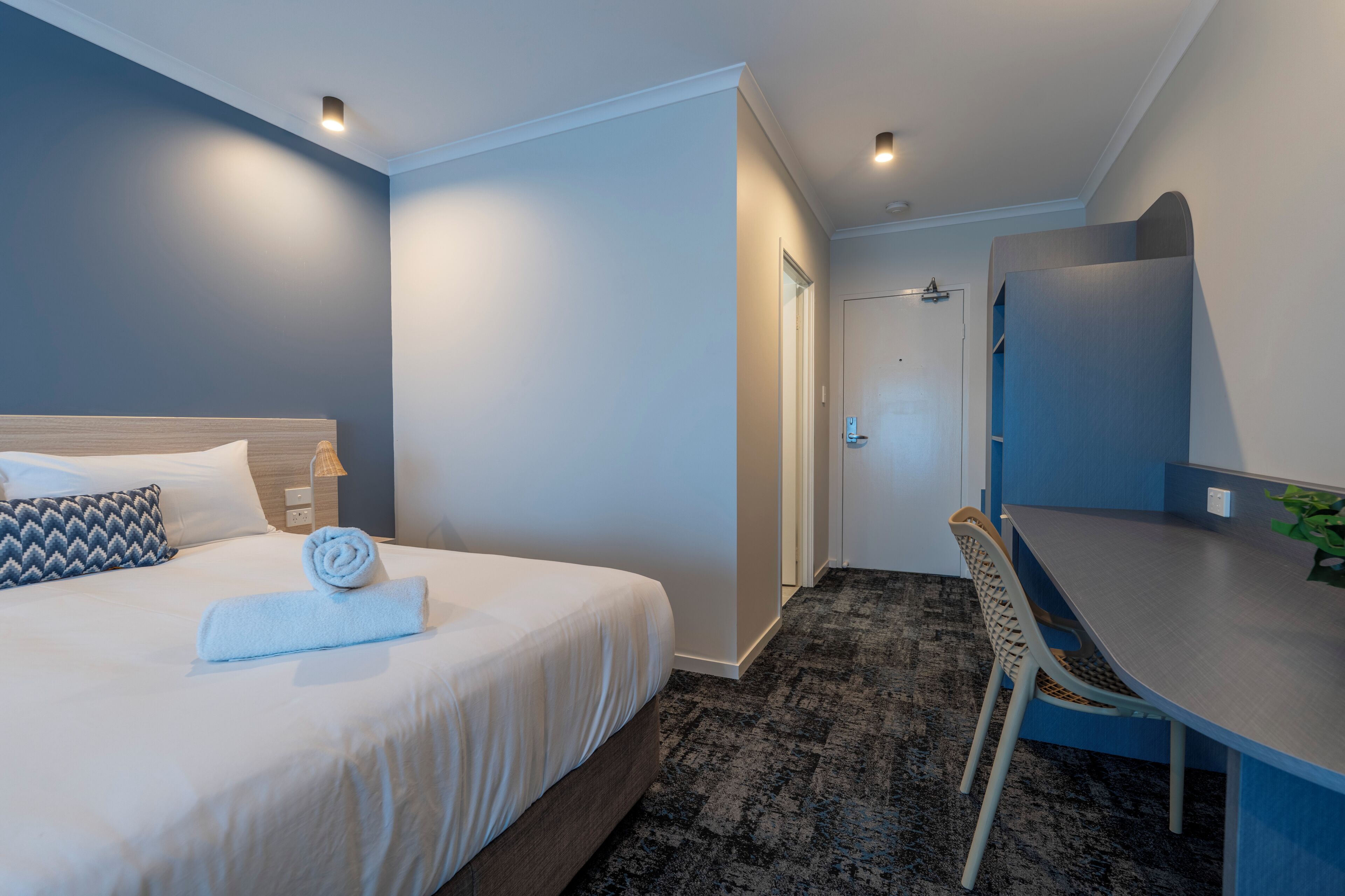 A hotel room with a bed, a desk, a chair, a door, and a wall-mounted mirror.