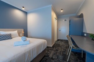 A hotel room with a bed, a desk, a chair, a door, and a wall-mounted mirror.