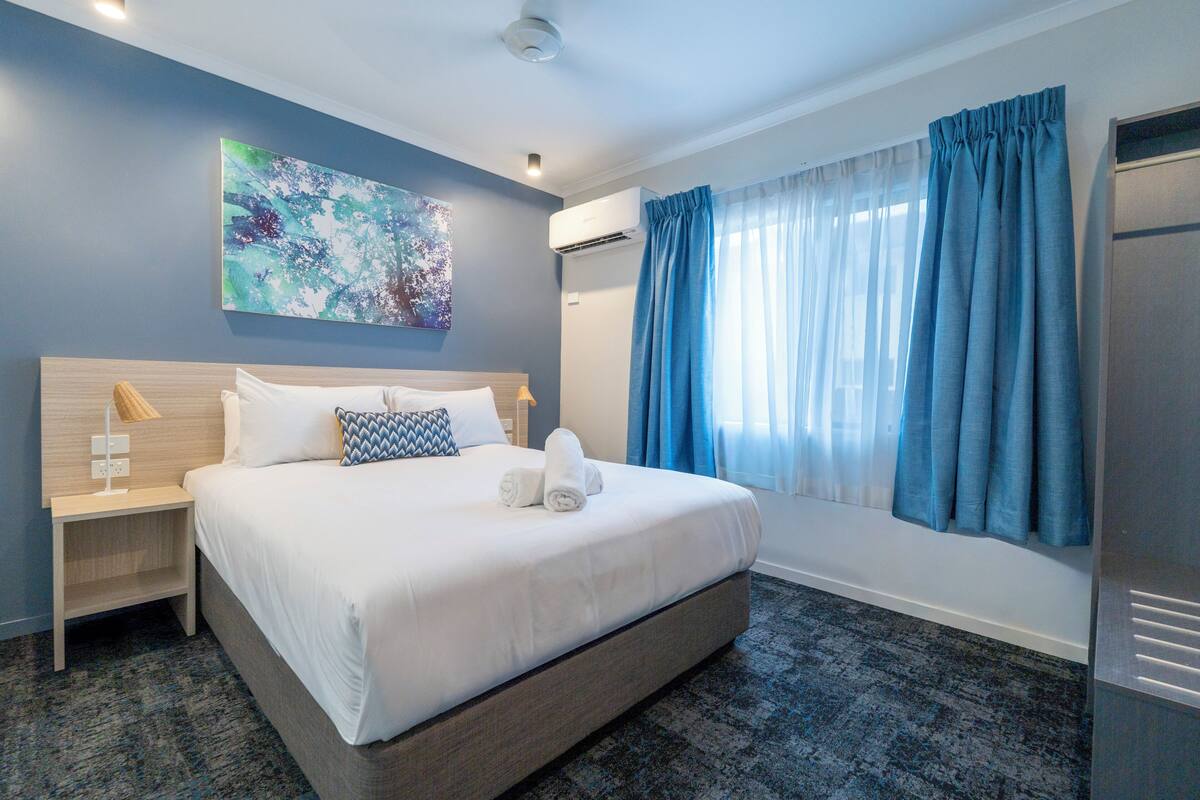 A hotel room with a bed, a nightstand, an air conditioning unit, a window with curtains, and a painting on the wall.