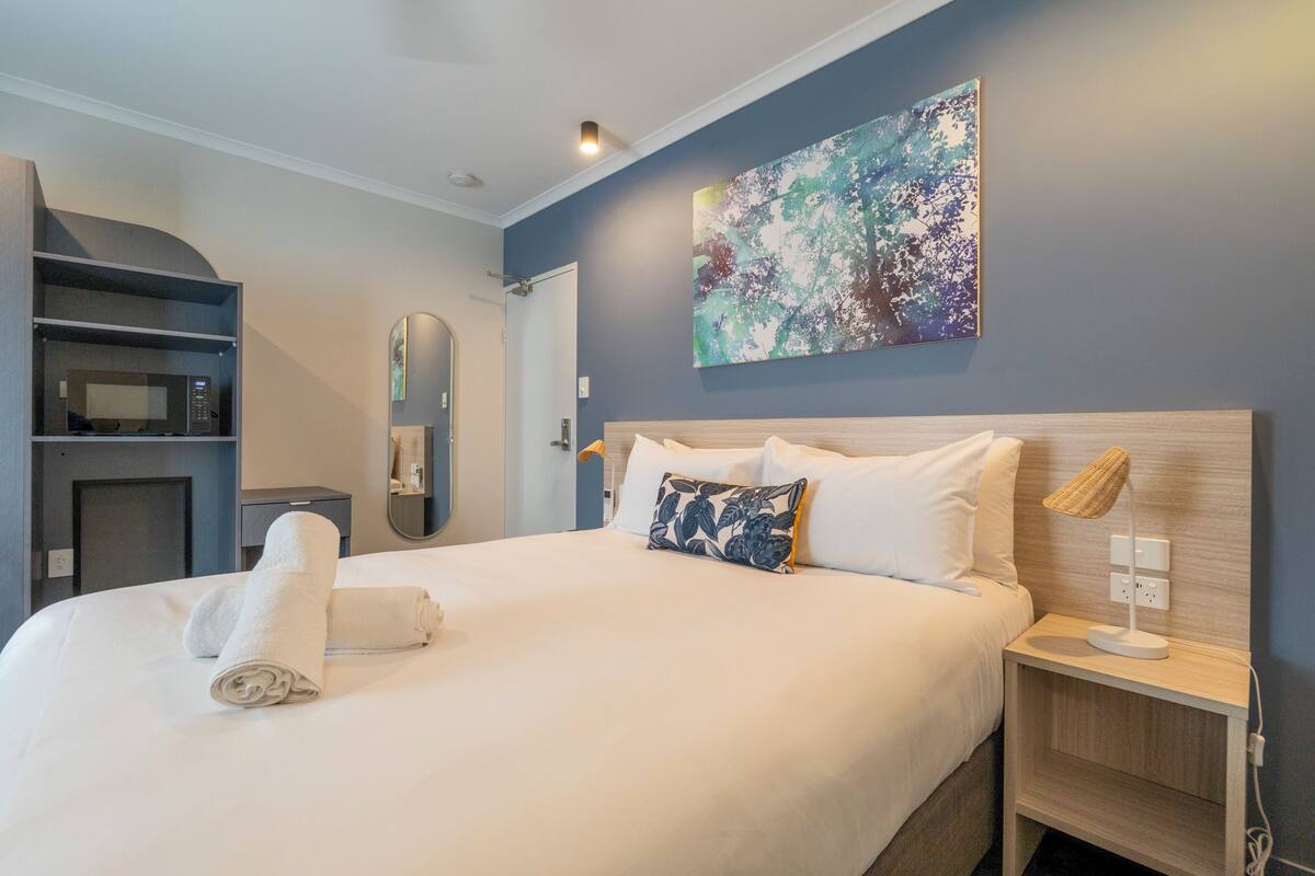 A hotel room with a bed, a bedside table with a lamp, a wall-mounted artwork, and a built-in microwave.