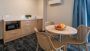 A compact kitchen with a microwave, a sink, and a potted plant. There is a round table with a fruit bowl and two wicker chairs.