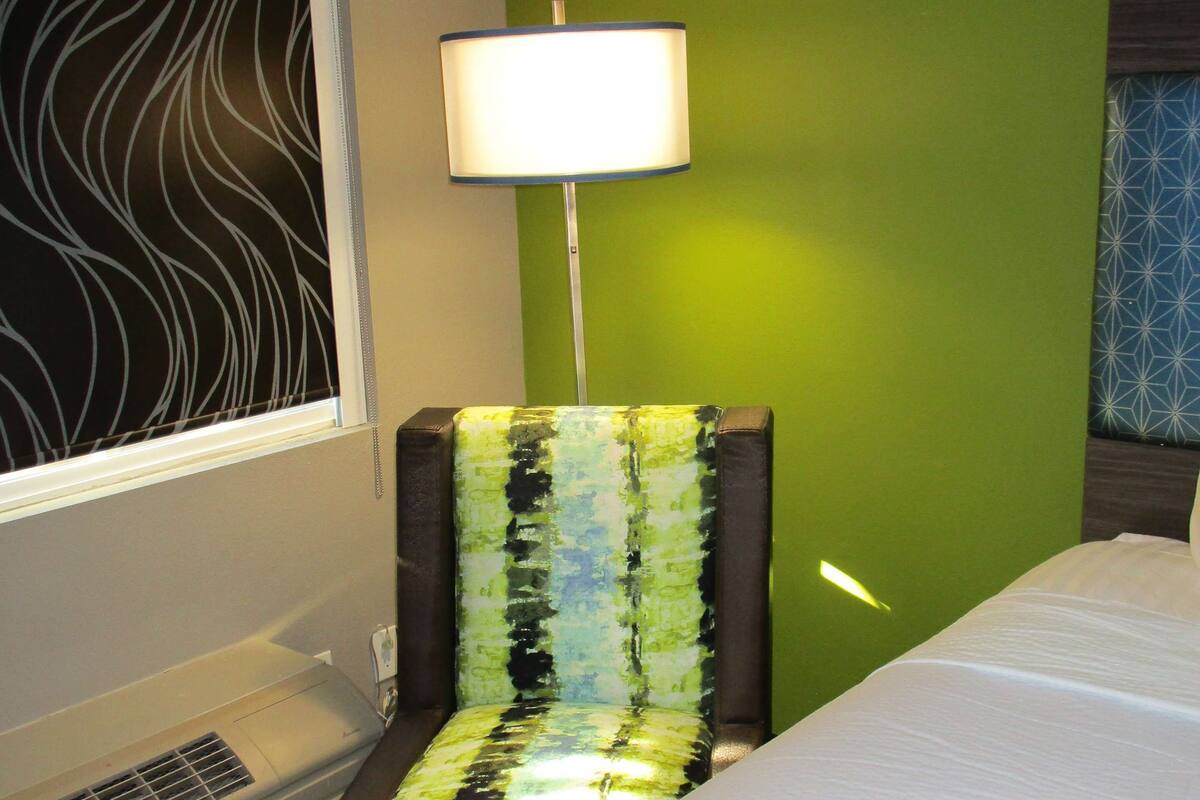 Ein Hotelzimmer mit einem Bett, einem Sessel mit farbenfroher Sitzauflage, einer Stehlampe, einem Fenster mit Schwarz-Weiß-Muster und einer Wand mit geometrischem Grün-Blau-Design.