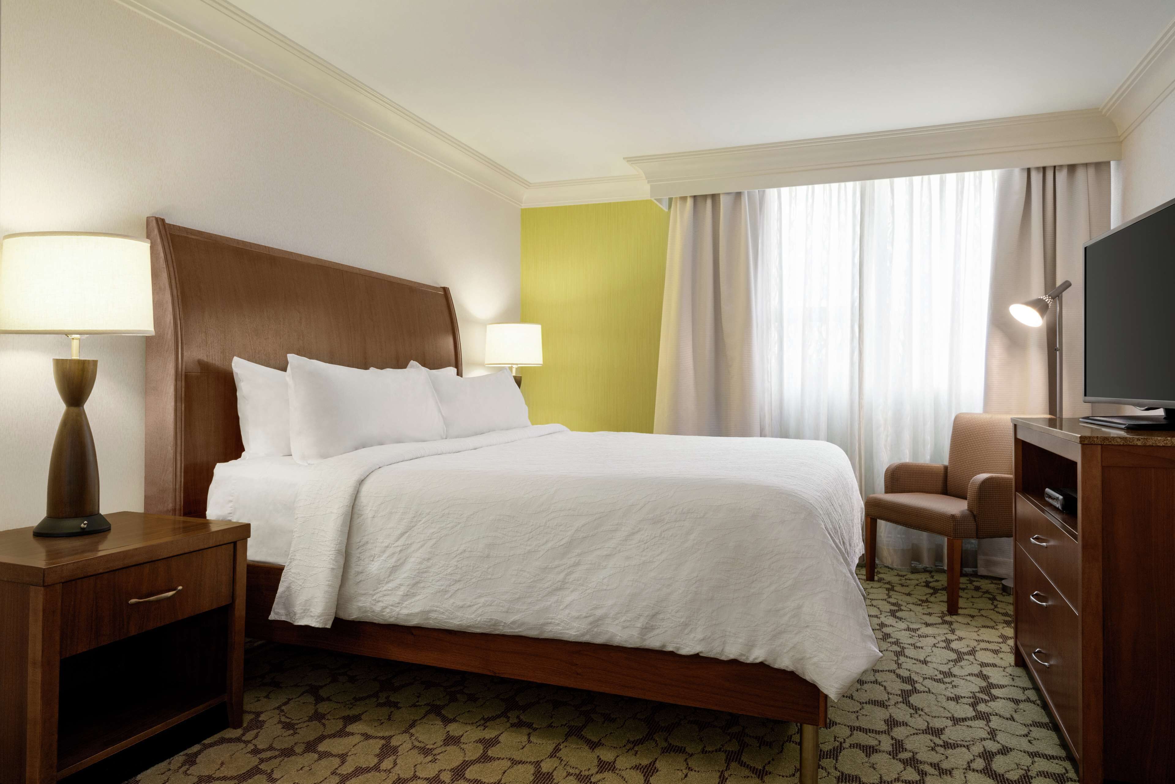 Une chambre d’hôtel avec un lit, des lampes de chevet, une chaise, une télévision et une fenêtre avec des rideaux.