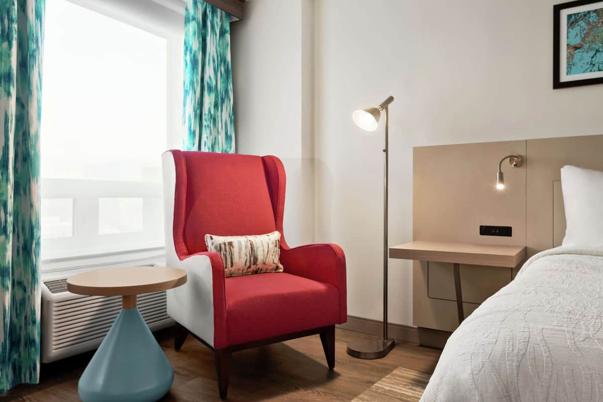 A hotel room with a red armchair, a bedside table, a floor lamp, and a bed.