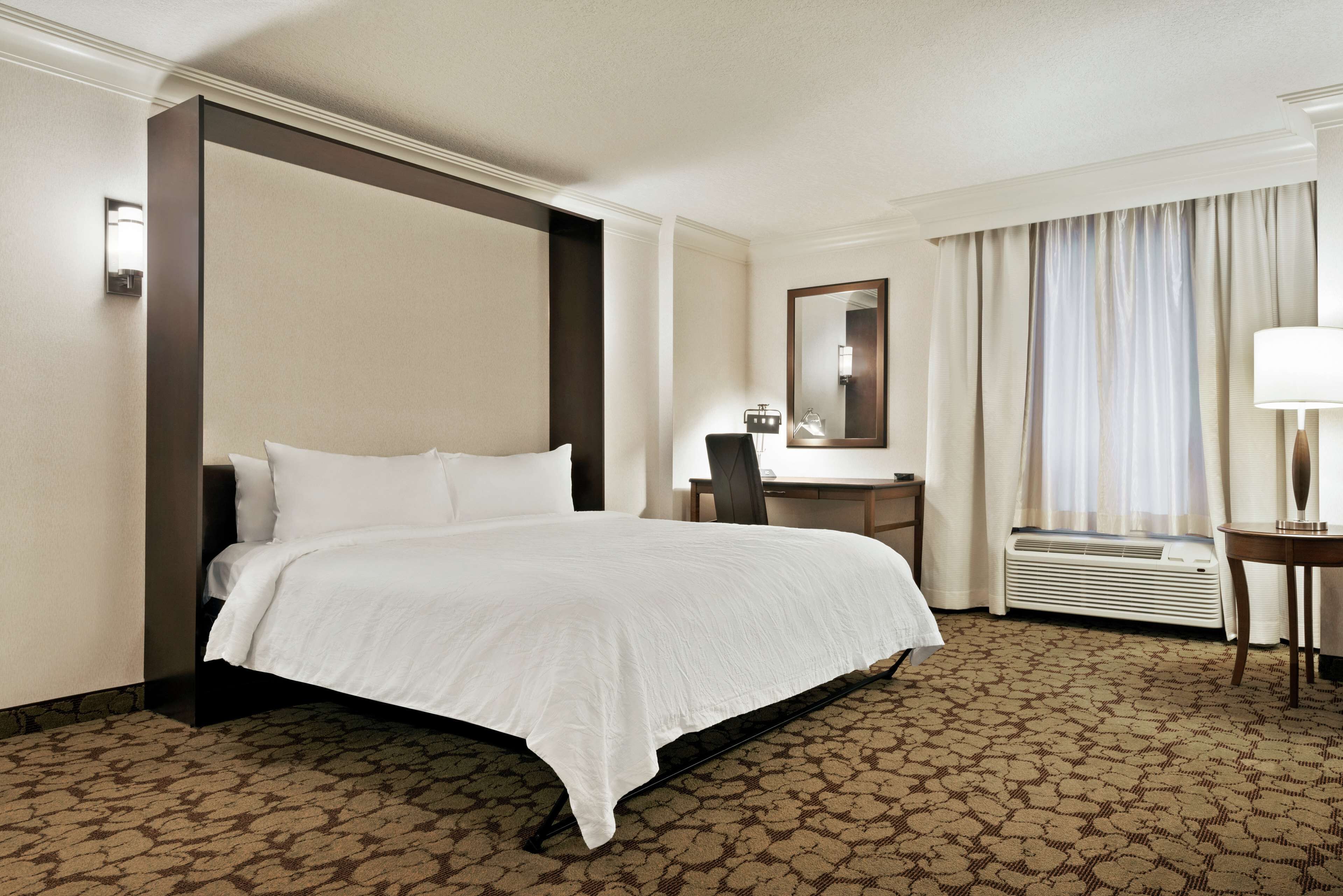 A hotel room with a large bed, a desk, a chair, a mirror, and an air conditioning unit.