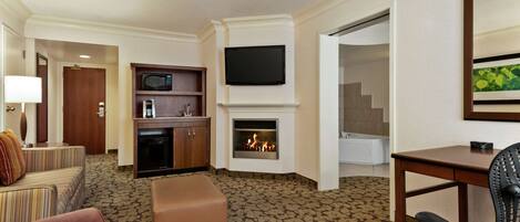 A hotel room with a living area featuring a sofa, ottoman, flat-screen TV, fireplace, and a dining table with a chair.