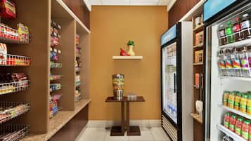 A grocery store interior with shelves stocked with various products, a refrigerated section, and a small table with a vase and salt shakers.