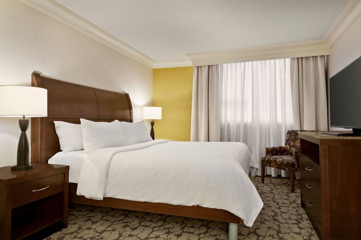 A hotel room with a bed, bedside tables, a TV, a chair, and a window with curtains.
