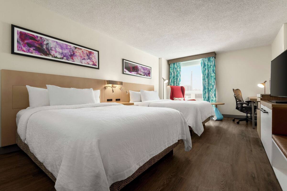 A hotel room with two beds, a desk, a chair, a TV, and artwork on the walls.