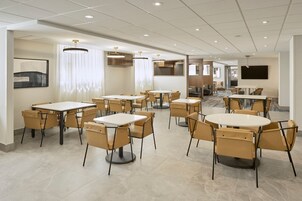 Comedor de hotel con varias mesas y sillas, luces empotradas en el techo y un televisor de pantalla plana grande montado en la pared.