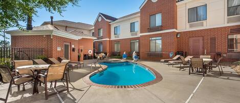 A poolside area with lounge chairs and tables, a brick building, and a clear blue sky.