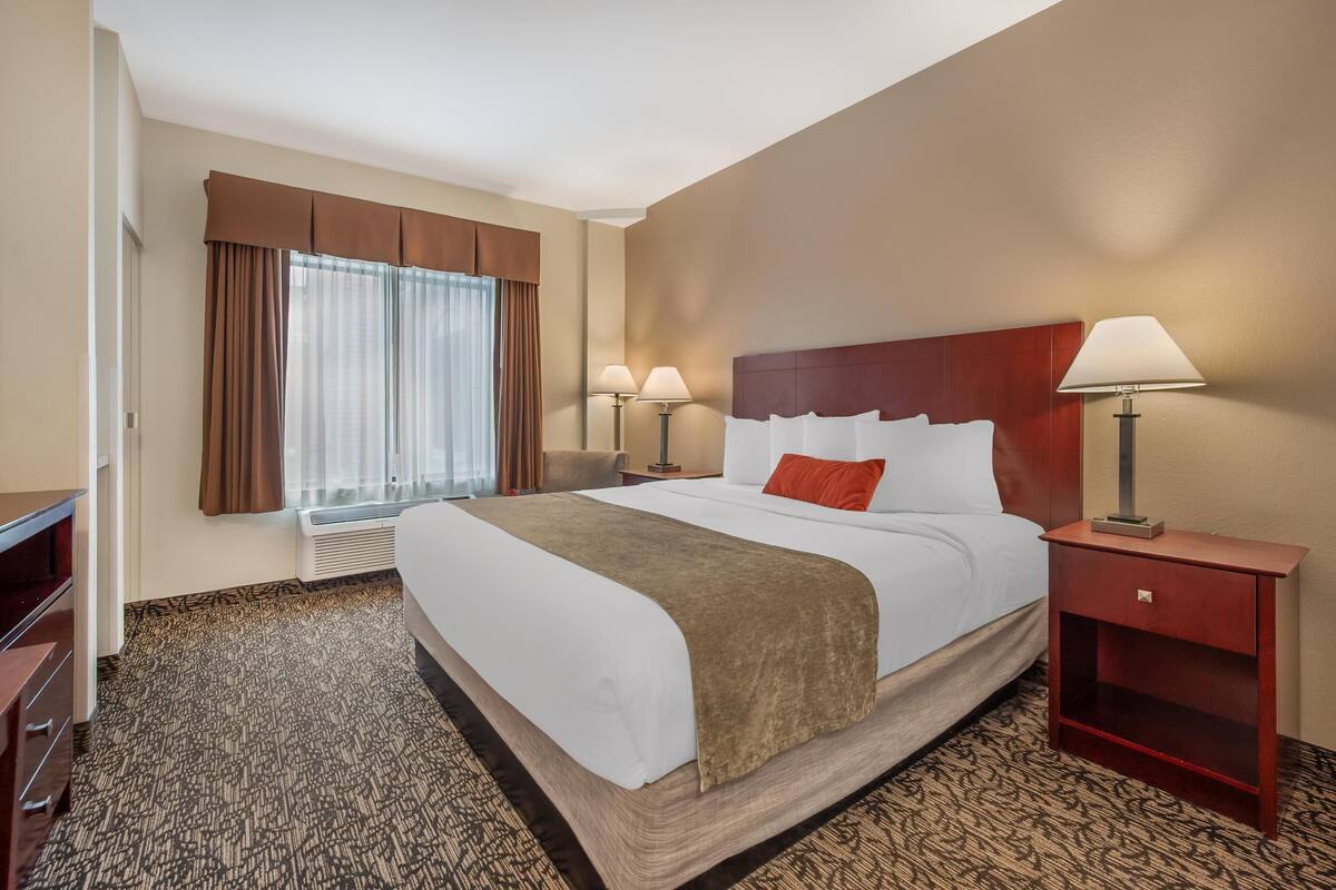 A hotel room with a large bed, two bedside lamps, a wooden nightstand, a desk, and a window with curtains.