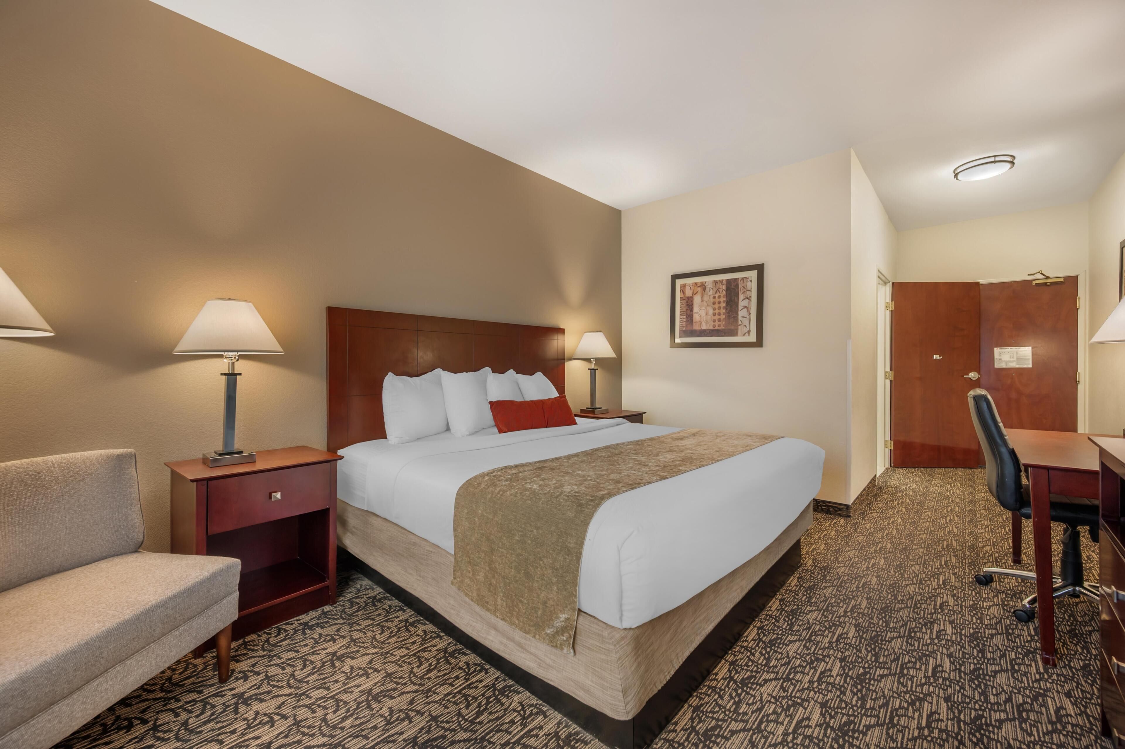 Quarto de hotel com uma cama grande, duas mesas de cabeceira com abajures, uma escrivaninha com cadeira e uma porta.
