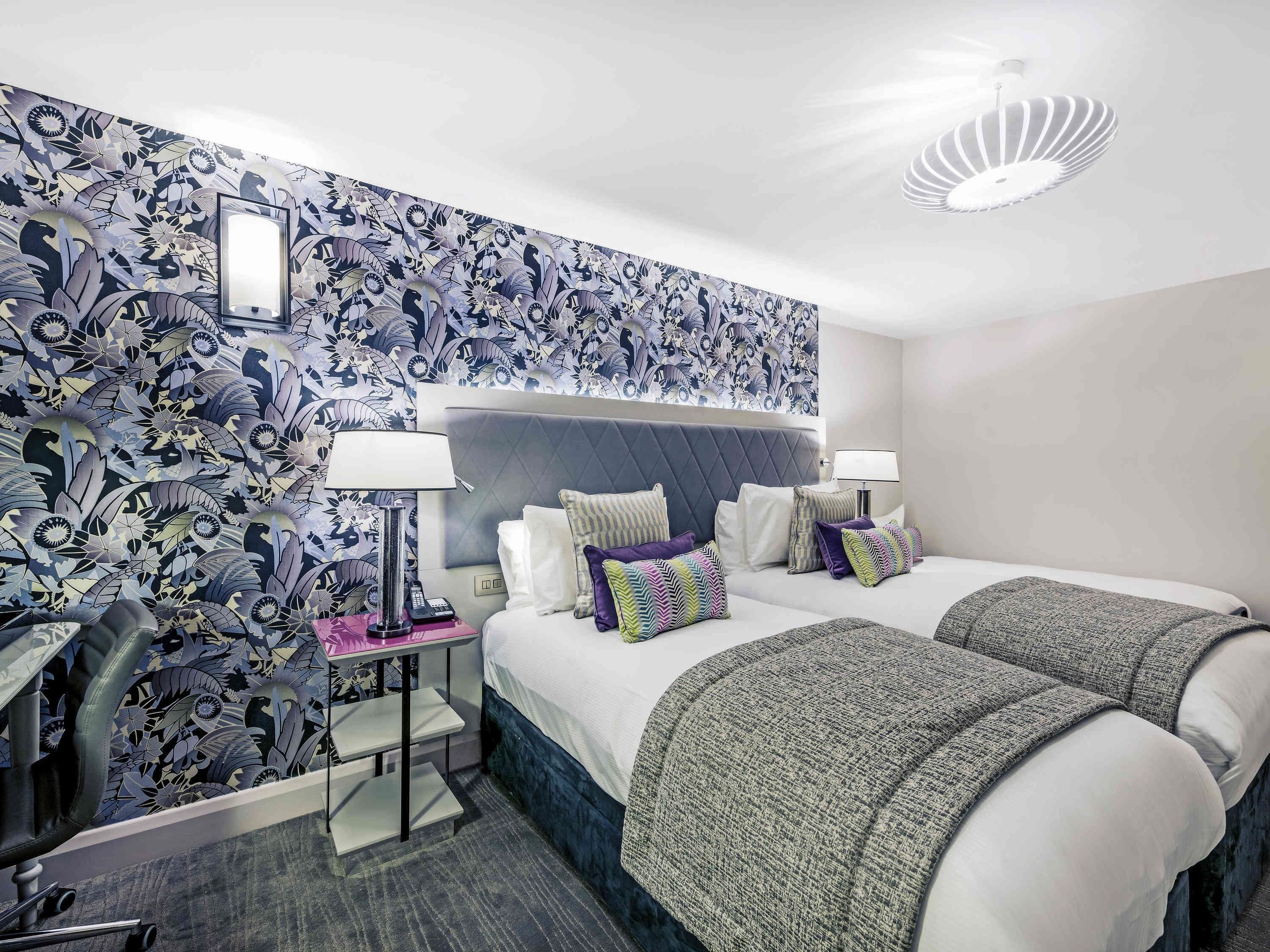 A hotel room with a patterned wallpaper, a bed with multiple pillows, a bedside table with a lamp, and a chair.