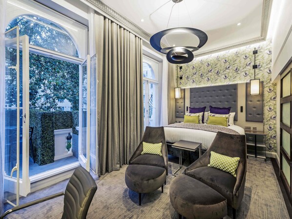 A modern hotel room with a large bed, a desk, two armchairs, and a view of trees through a large window. - Mercure London Hyde Park Hotel (London)