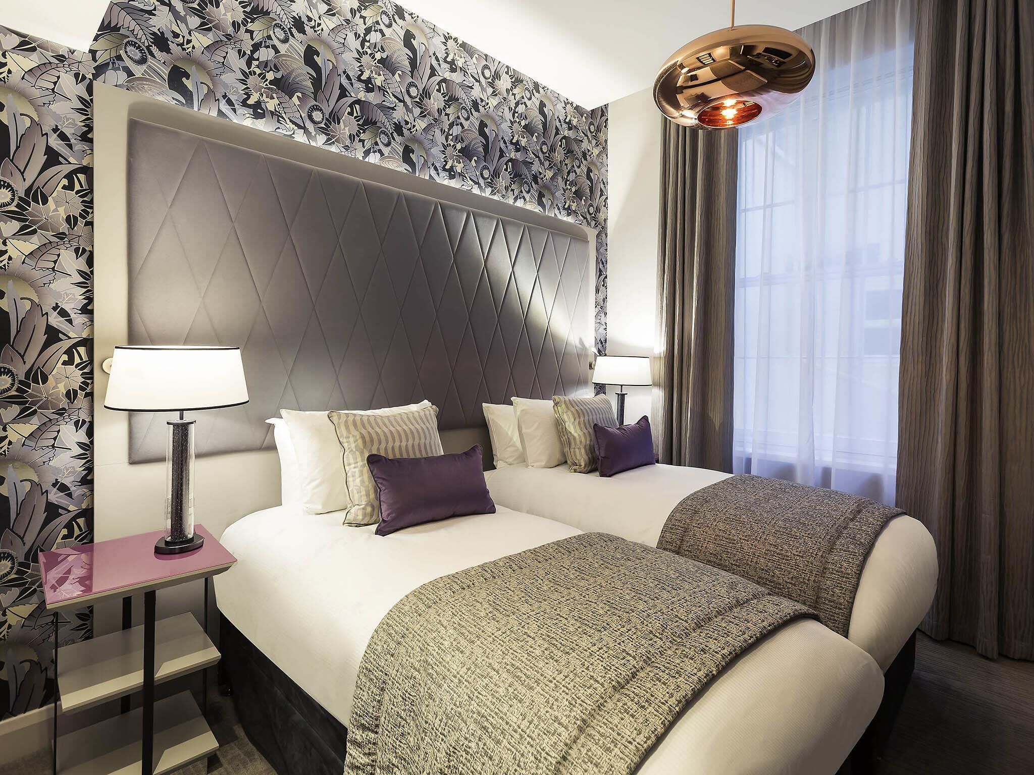 A hotel room with a large bed, two bedside tables, a modern chandelier, and a patterned wallpaper.