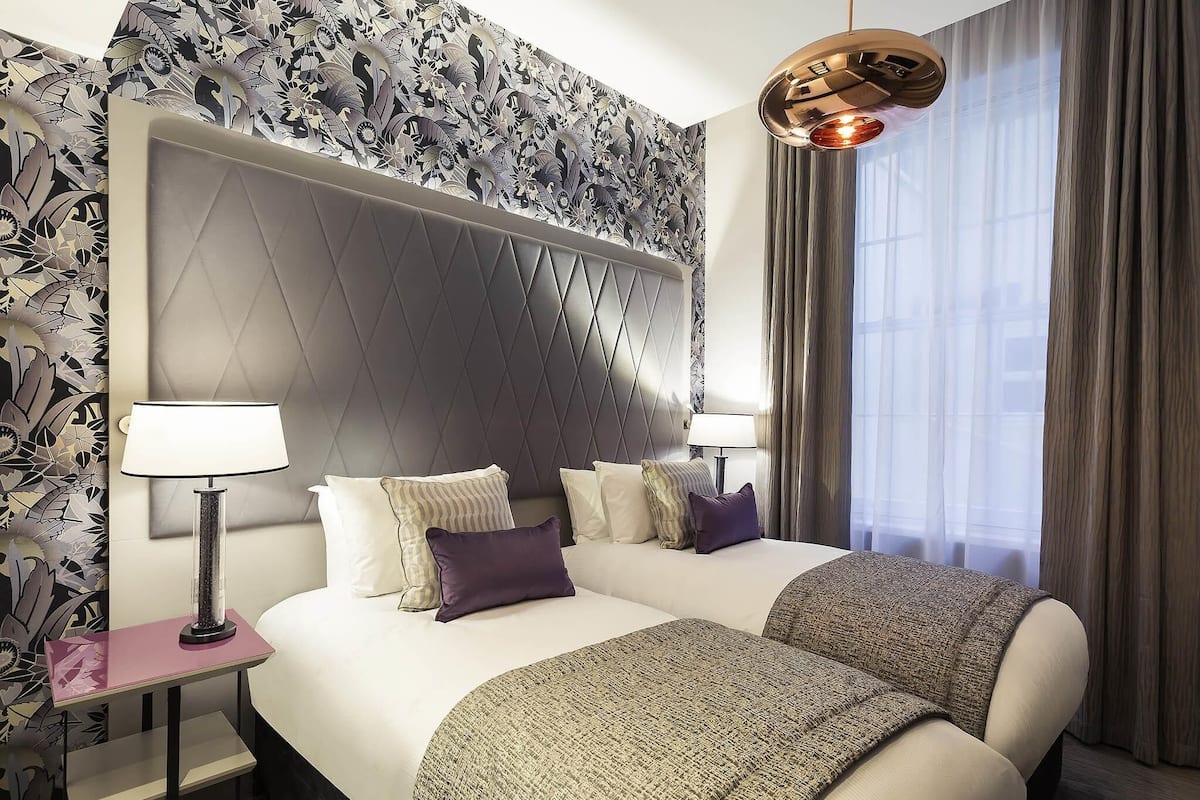 Une chambre d’hôtel avec un grand lit, deux tables de chevet, un lustre moderne et un papier peint à motifs.