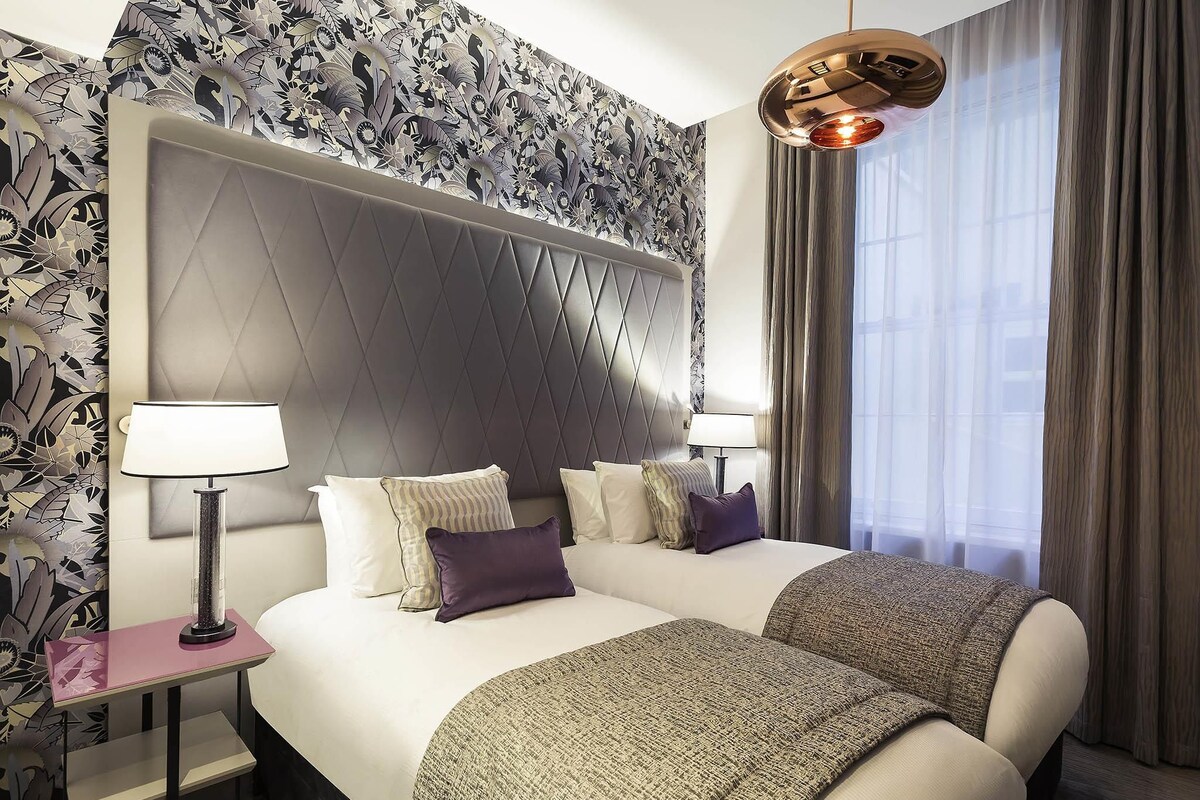 Een hotelkamer met een groot bed, twee nachtkastjes, een moderne kroonluchter en behang met een patroon.