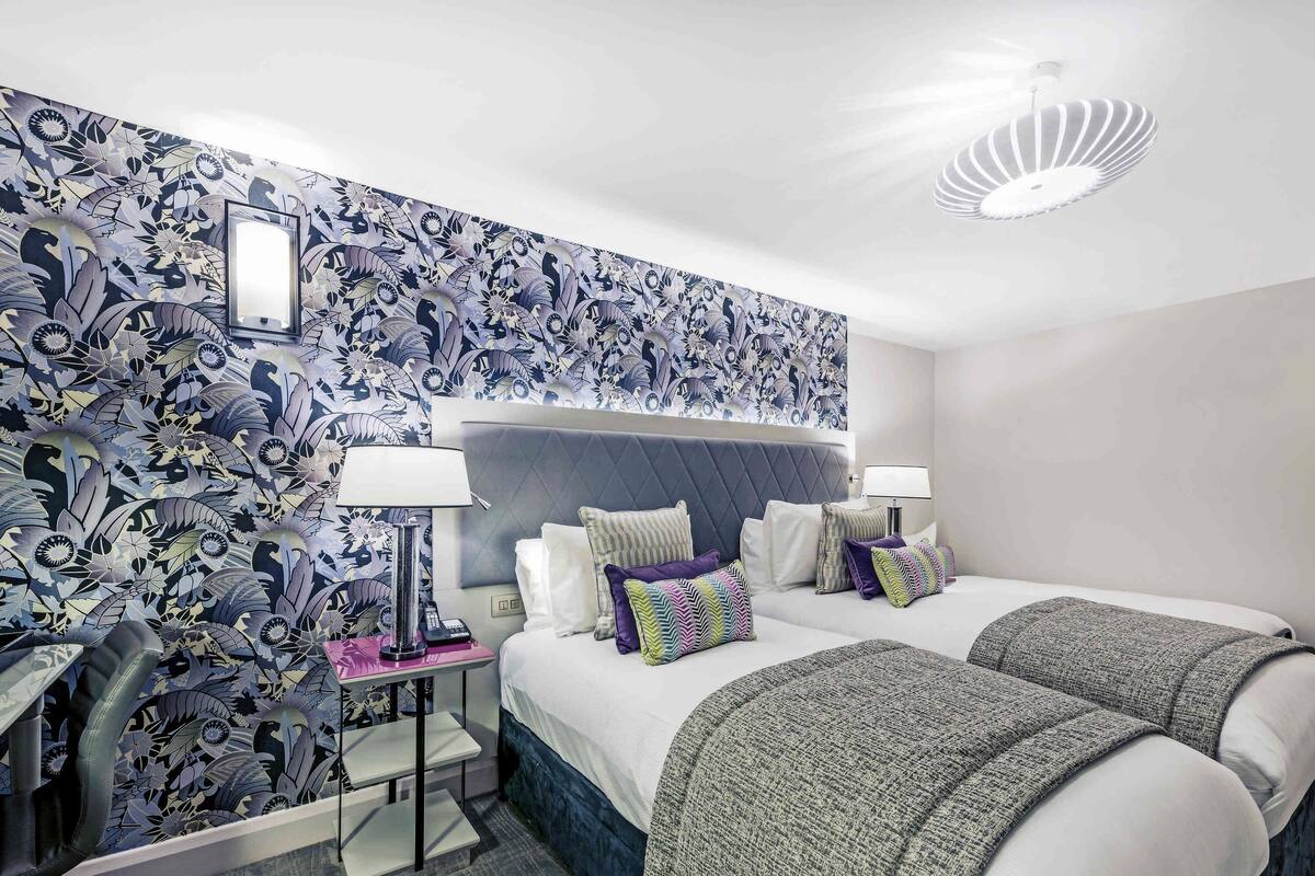 Ein Hotelzimmer mit strukturiertem Wandpapier, einem Bett mit vielen Kissen, einem Nachttisch mit Lampe und einem Sessel.