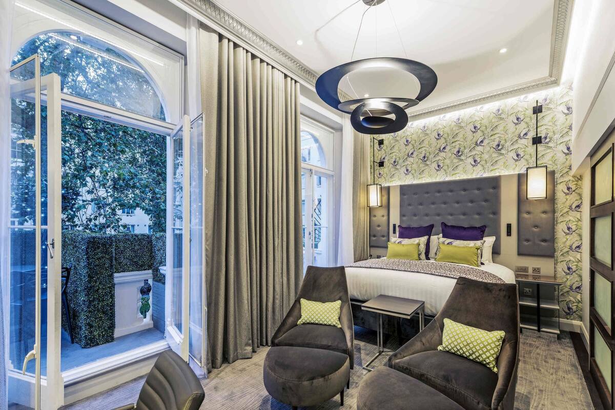Ein modernes Hotelzimmer mit einem großen Bett, einem Schreibtisch, zwei Sesseln und Blick auf Bäume durch ein großes Fenster.