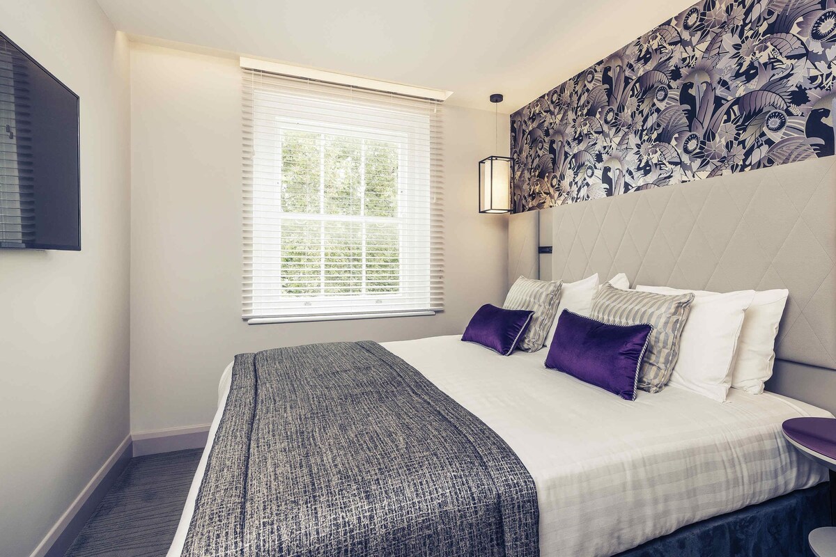 Een moderne hotelkamer met een groot bed, een hoofdbord met patroon, een raam met zonwering en een aan de muur bevestigd kunstwerk.