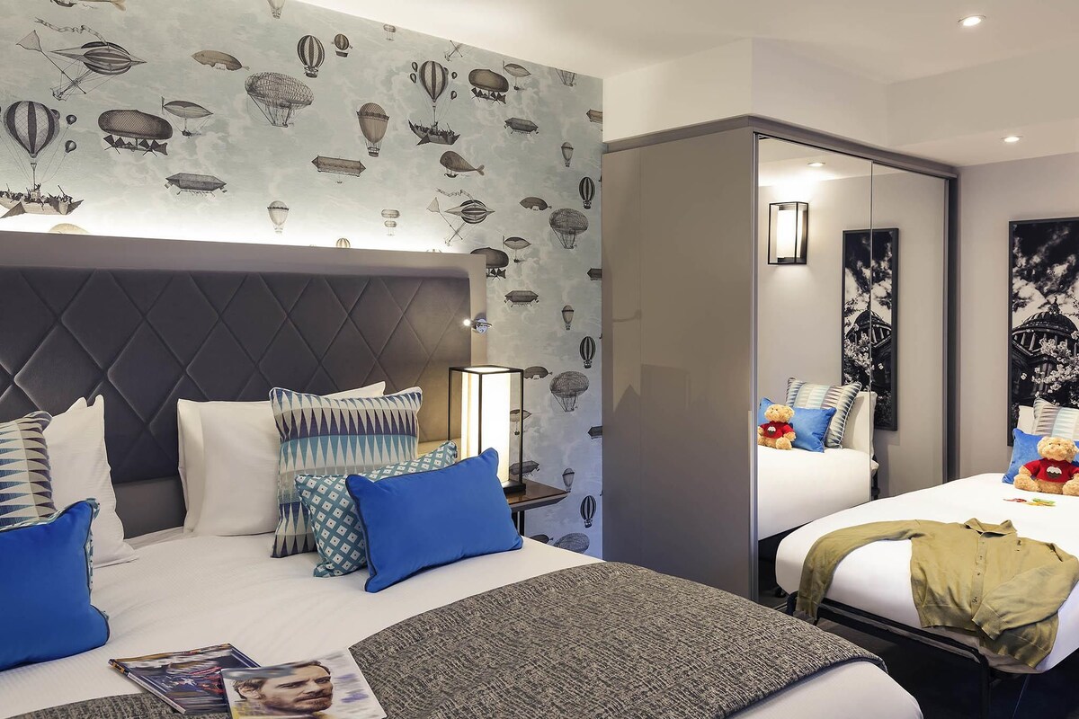 Een hotelkamer met een groot bed, een kleiner bed, een nachtkastje, een lamp en een muur met een patroon van luchtballonnen.