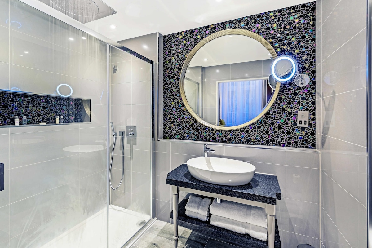 A modern bathroom with a glass-enclosed shower, a round mirror with a light, and a black countertop with a sink.