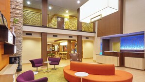 Eine geräumige Lobby mit hoher Decke, einem Empfangsbereich und einer Sitzecke mit violetten und orangen Stühlen.