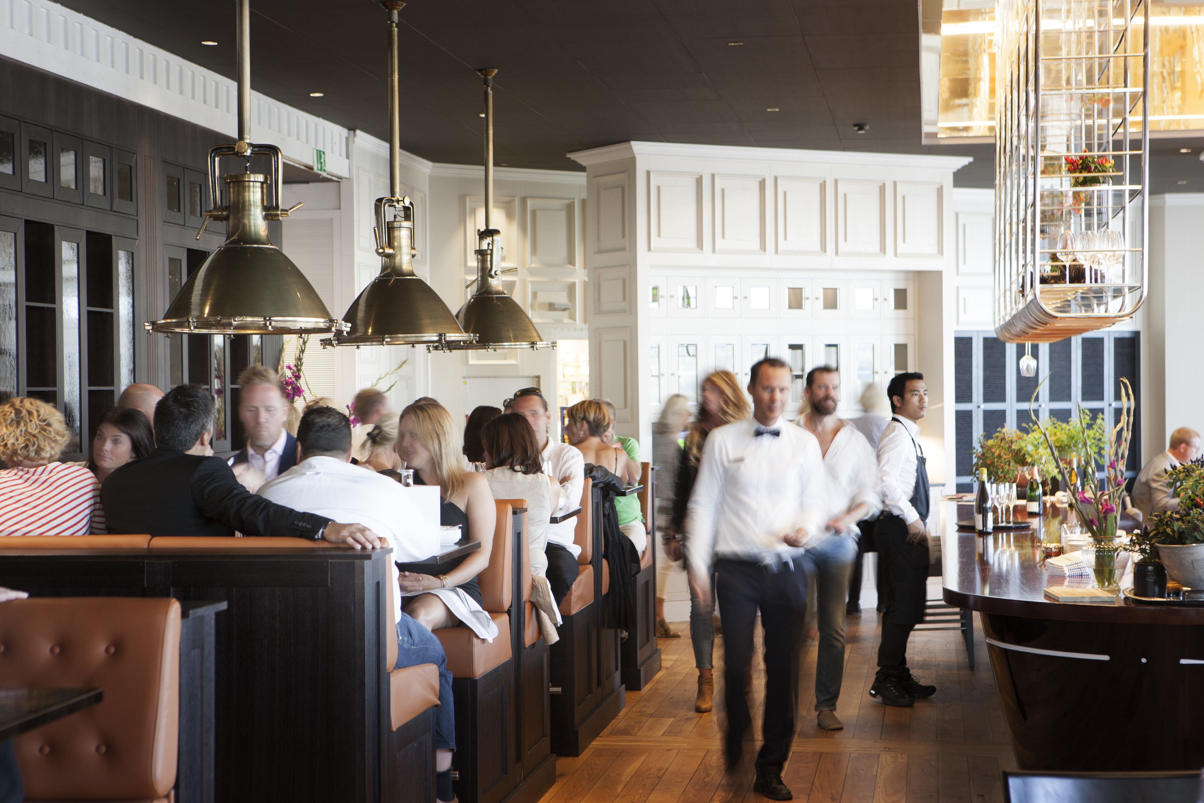 Un restaurant avec des banquettes en bois, des luminaires suspendus et un coin bar agrémenté d’un vase de fleurs.