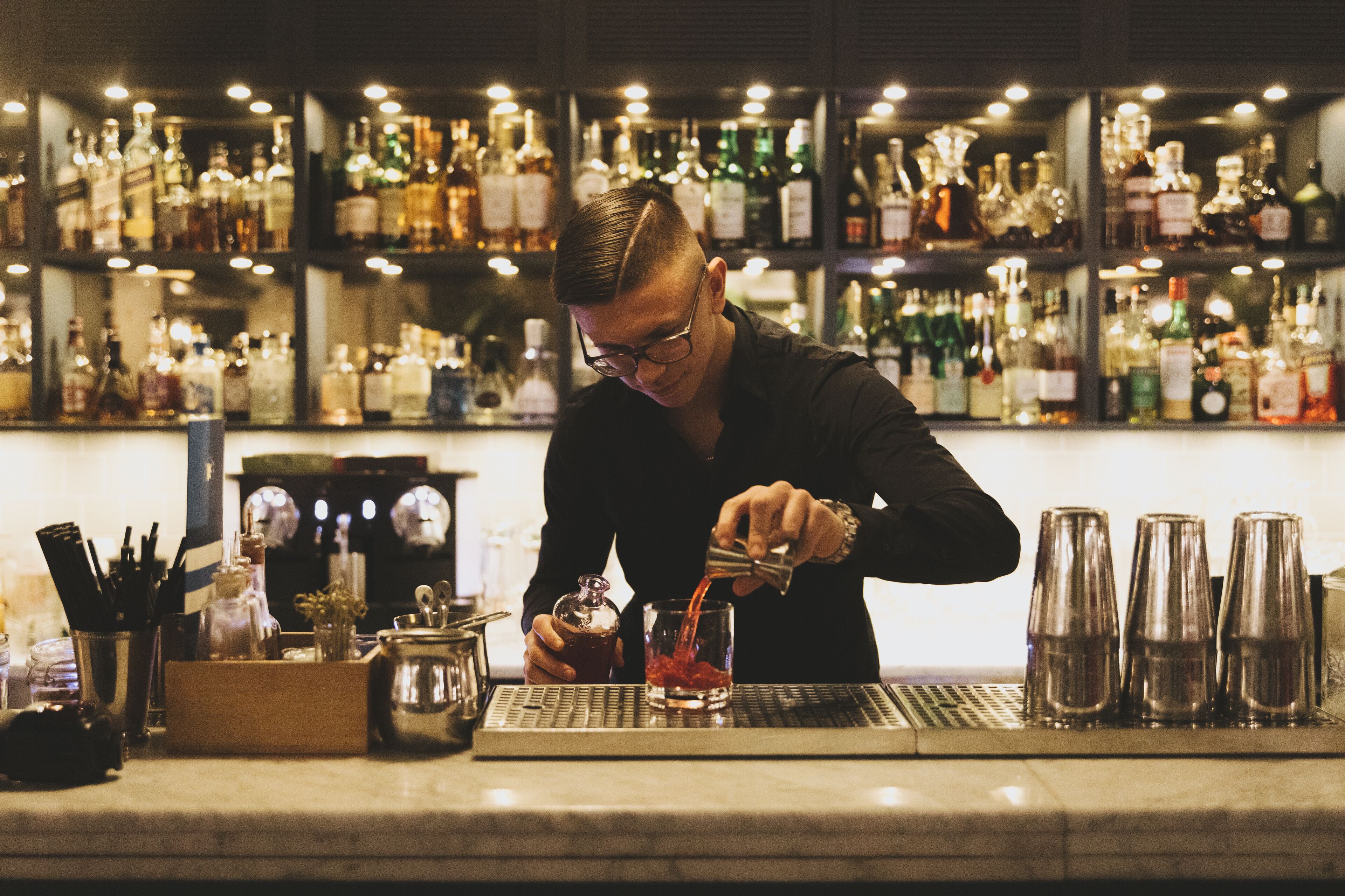 A bartender pouring a drink at a bar with shelves of liquor bottles in the background.
