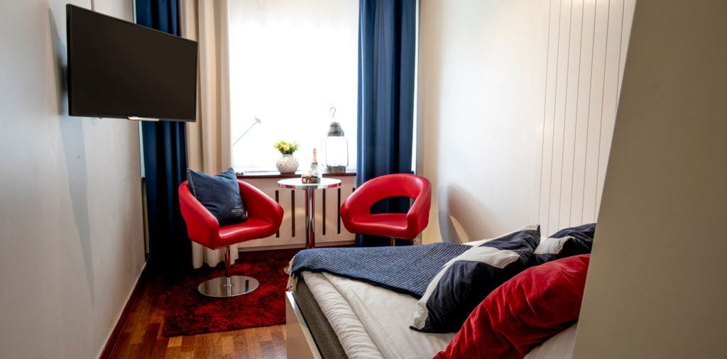 Hotellihuone, jossa on sänky, kaksi punaista tuolia, pieni pöytä lampun kanssa ja seinälle asennettu televisio.