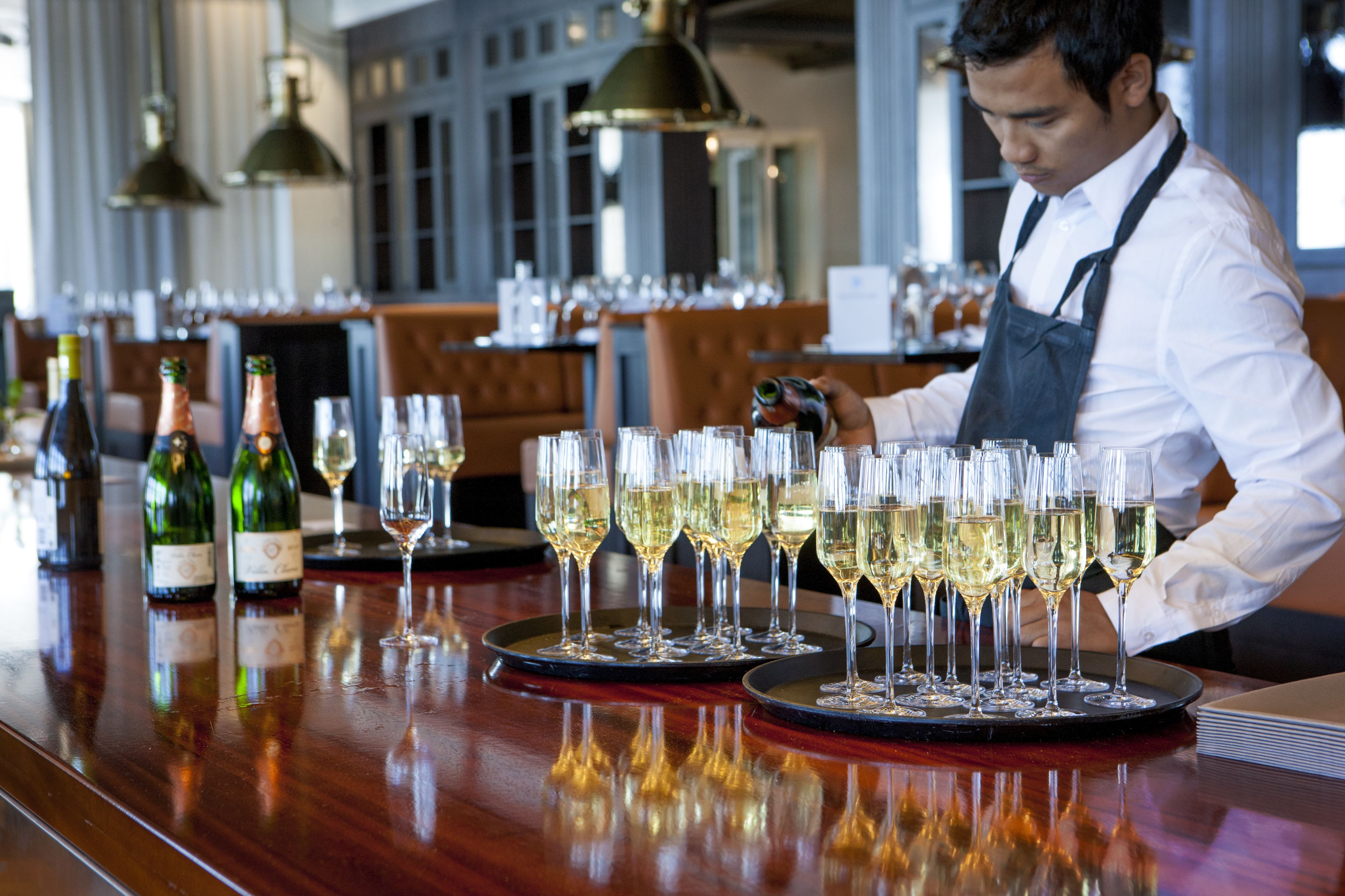A bar with multiple glasses of white wine on trays, a bartender, and bottles of wine on the counter.