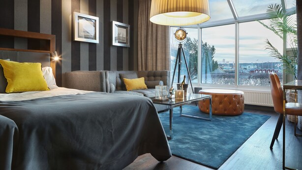 Una habitación de hotel moderna con una cama grande, un sofá, un reposapiés redondo, una mesita de centro y un ventanal con vistas a la ciudad.