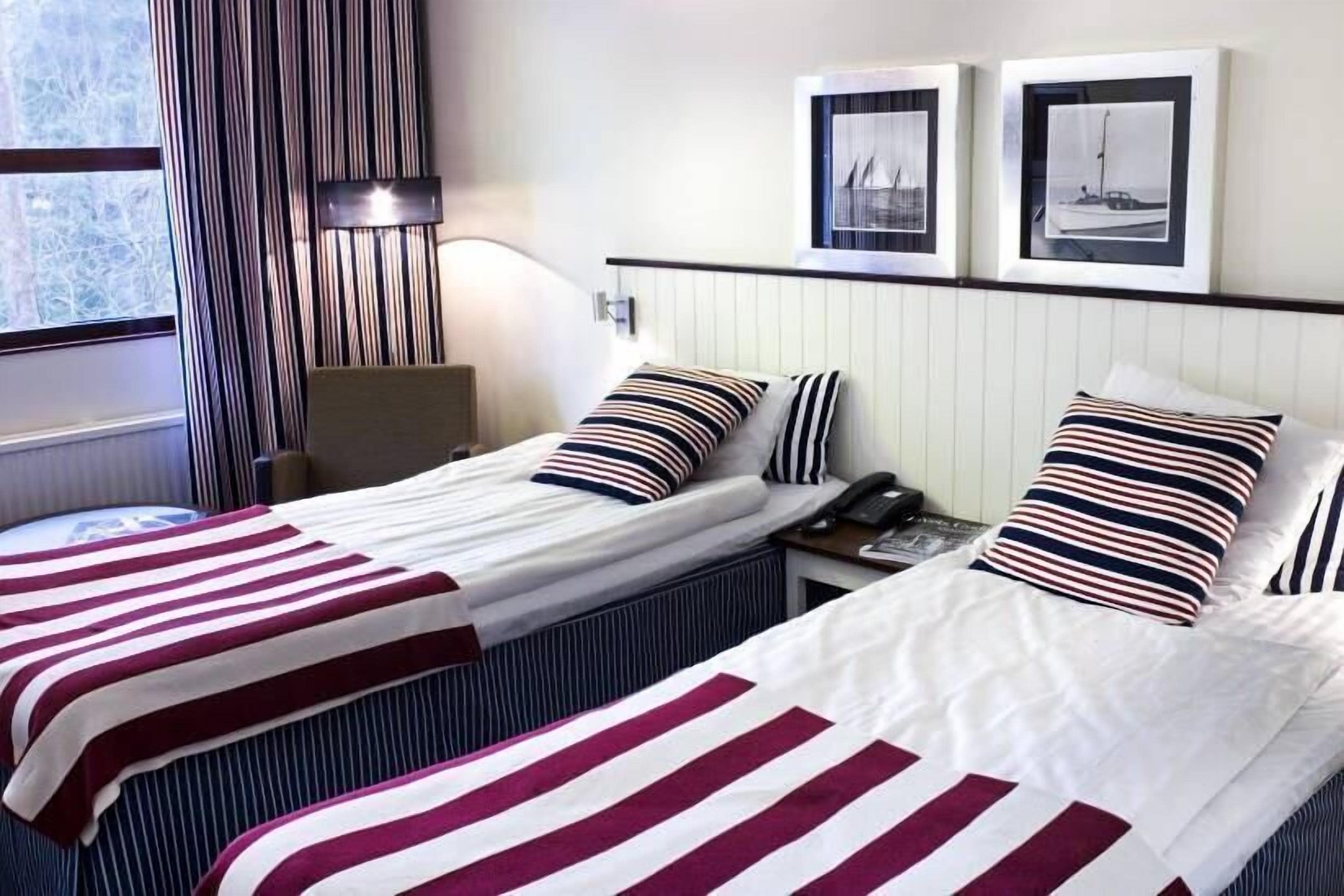 Habitación de hotel con dos camas, ropa de cama a rayas, una mesita de noche con teléfono, lámpara y cuadros en la pared.