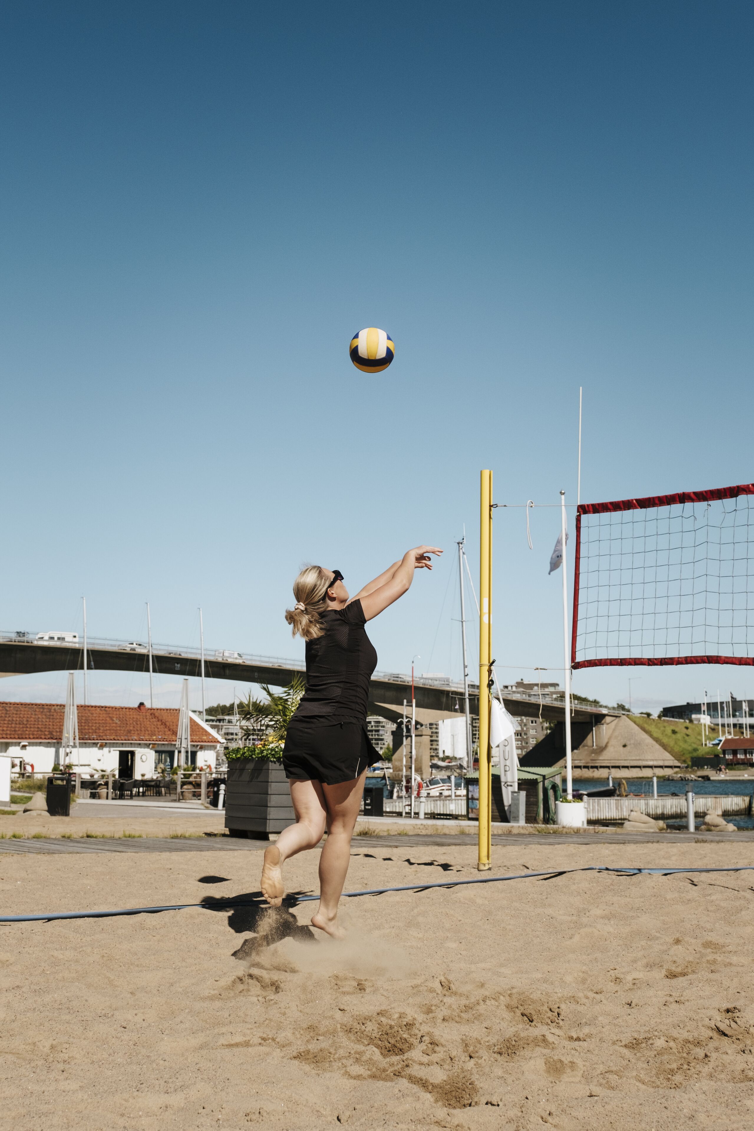 A person playing volleyball on a sandy court with a net, buildings, and a bridge in the background.