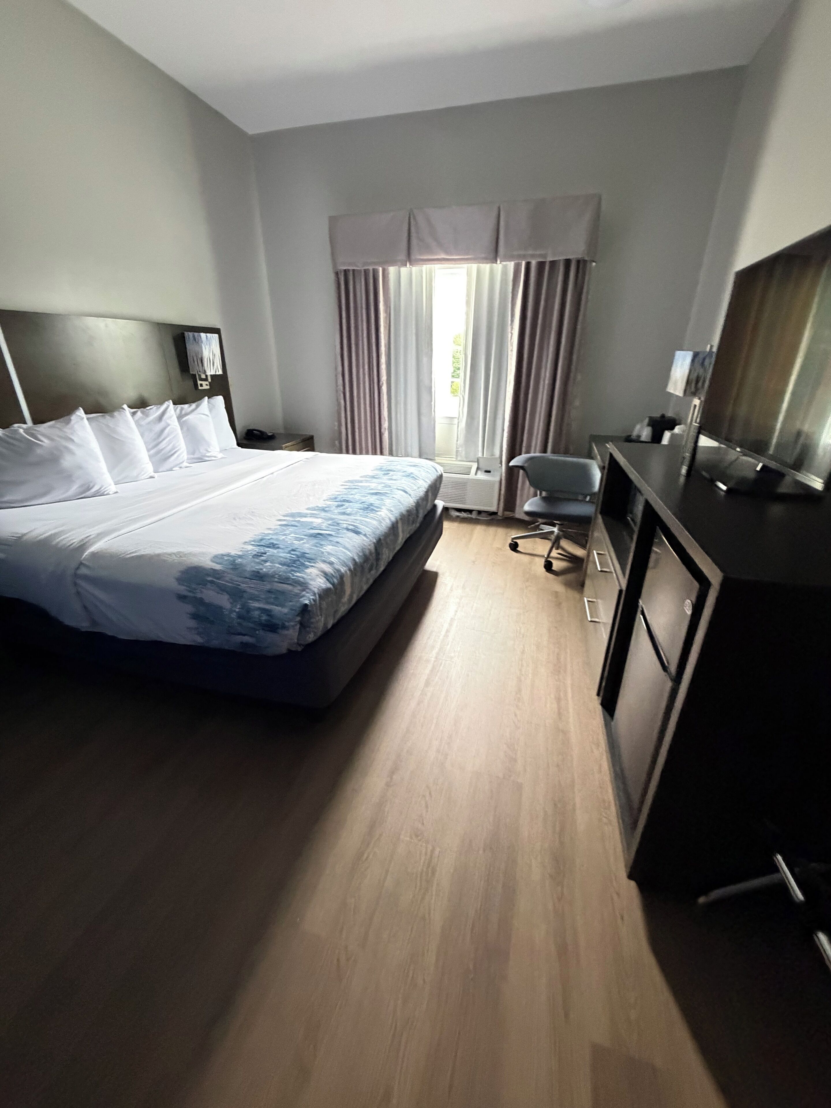 A hotel room with a large bed, a desk with a chair, a television, and a window with curtains.