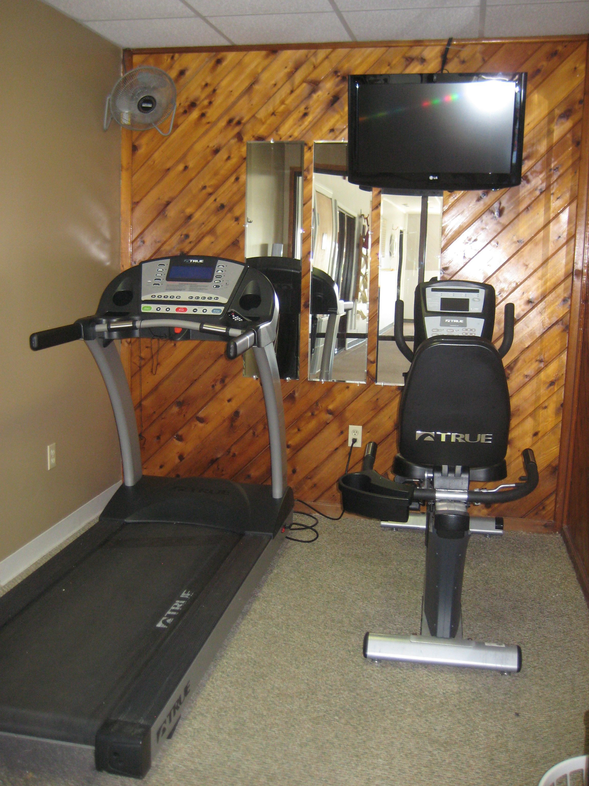 Un gimnasio en casa con una cinta de correr, una bicicleta estática y un televisor montado en la pared.