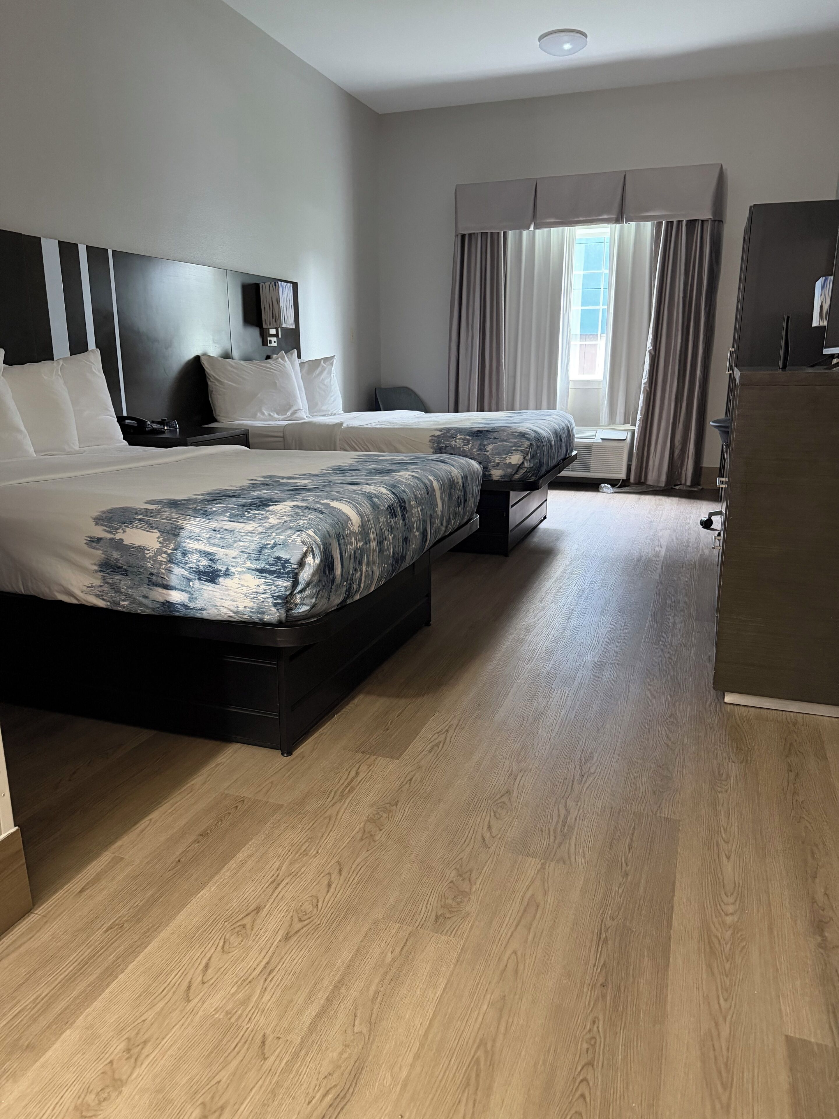 A modern hotel room with two beds, a flat-screen TV, a desk, and a window with curtains.