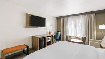 A hotel room with a bed, a desk with a computer, a chair, a television mounted on the wall, and a window with curtains.