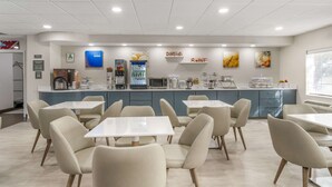 A modern cafe with white tables and chairs, a counter with coffee machines, and artwork on the walls. - Comfort Inn & Suites Louisville Airport Fair & Expo (Louisville)