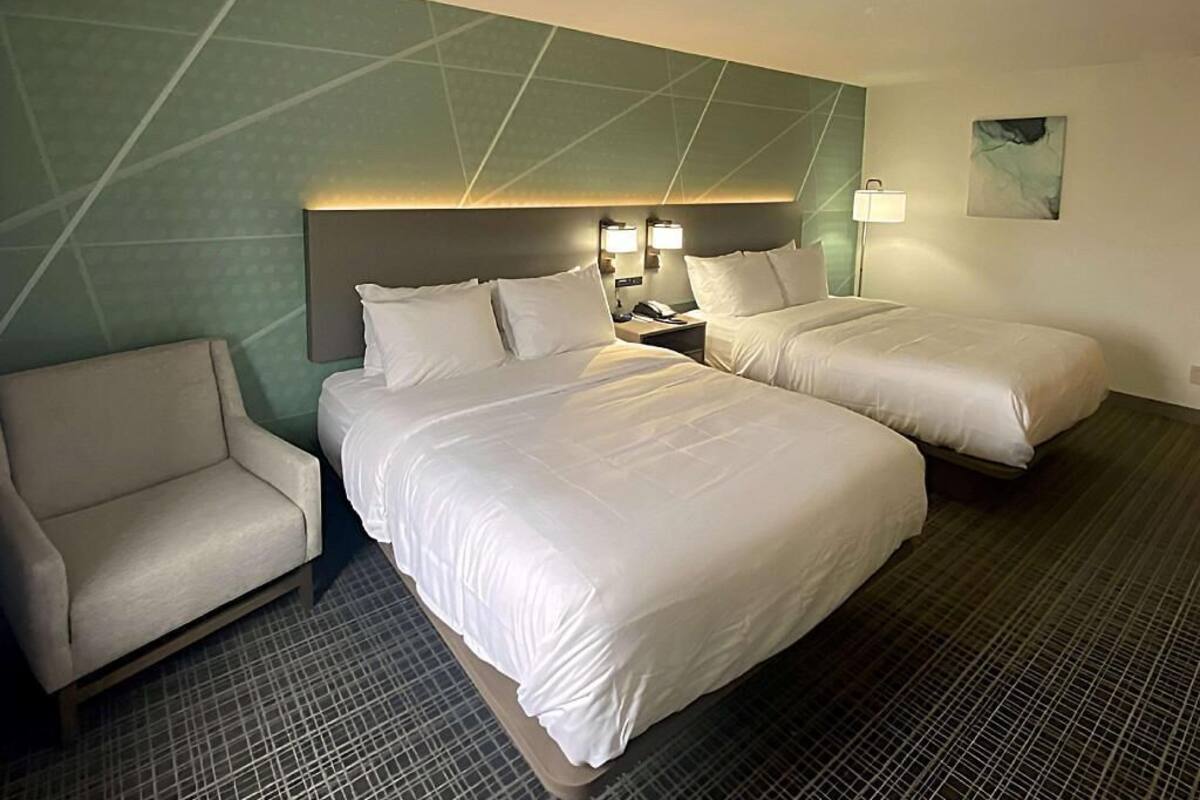 A hotel room with two beds, a sofa, a headboard with integrated lighting, a nightstand, and a wall-mounted lamp.