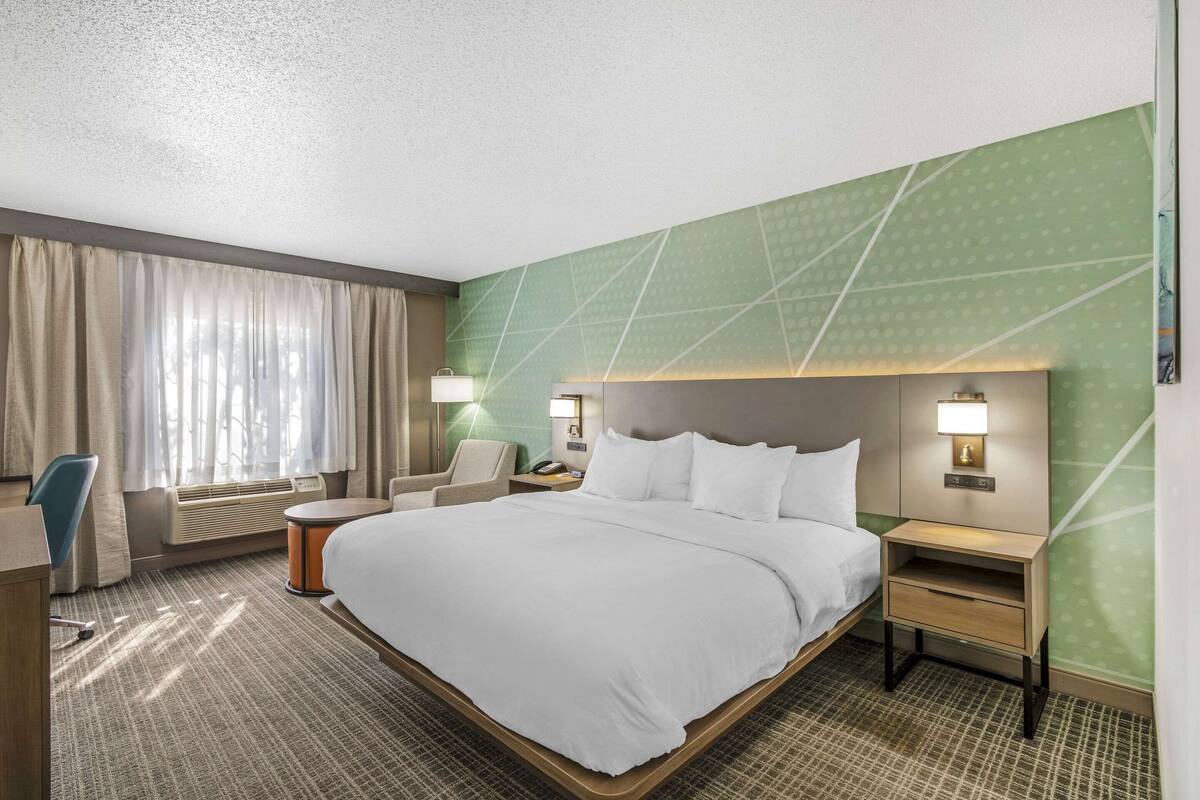 A hotel room with a large bed, a desk, a chair, a nightstand, and a wall-mounted lamp.