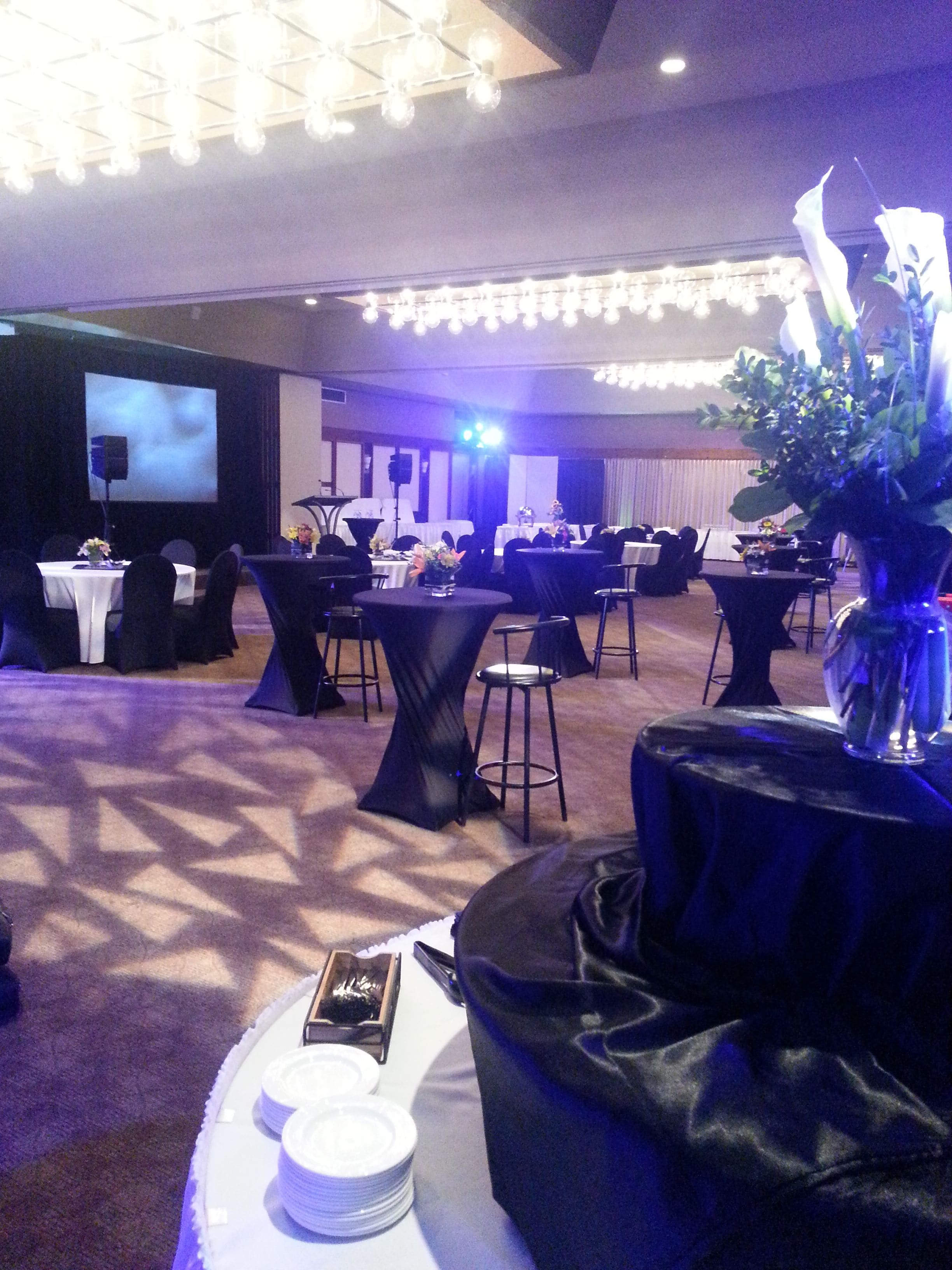 Une salle de banquet avec des tables rondes, des chaises et une scène équipée d’un écran.