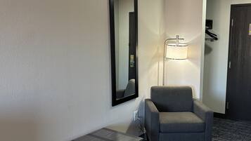 A hotel room with a grey armchair, a small table, a mirror, and a floor lamp.