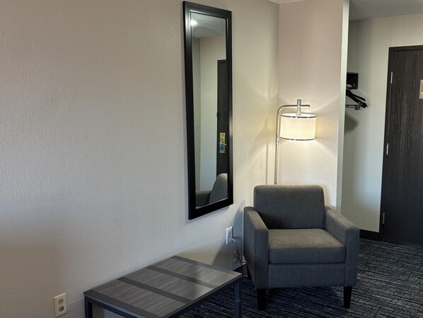 A hotel room with a grey armchair, a small table, a mirror, and a floor lamp.