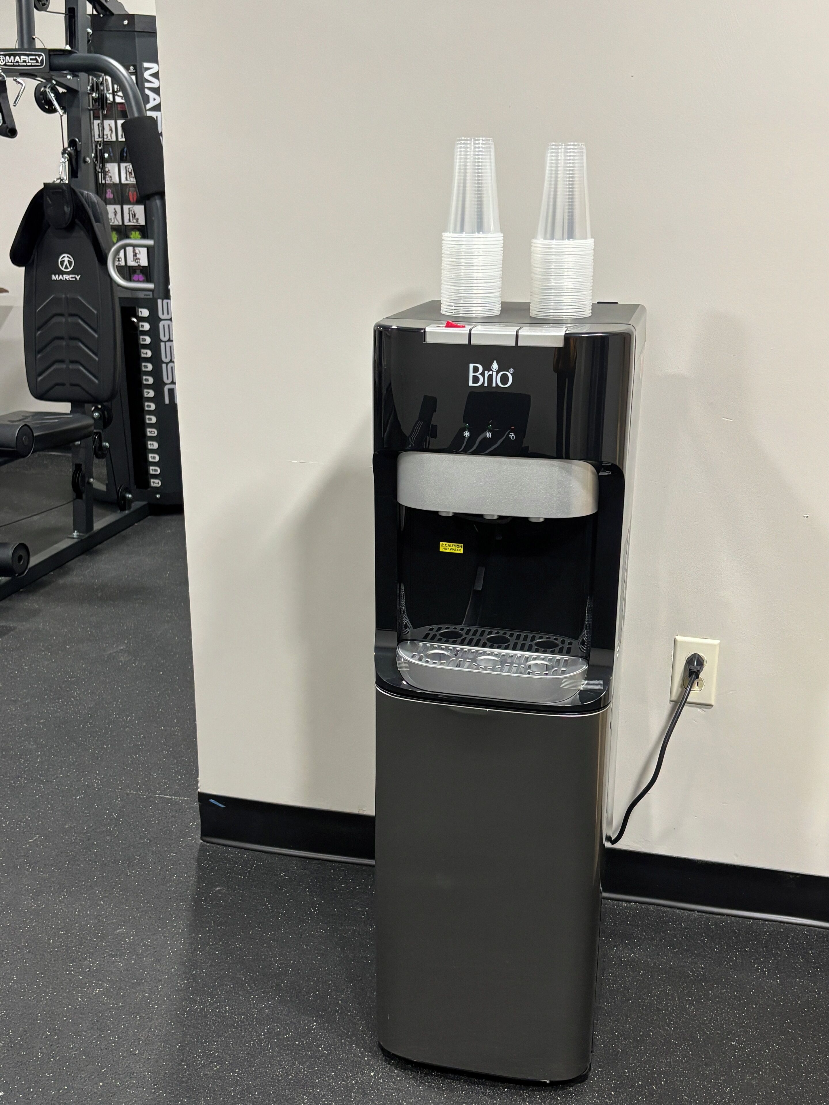 A water dispenser with two cups on top, a gym machine in the background, and a power outlet on the wall.