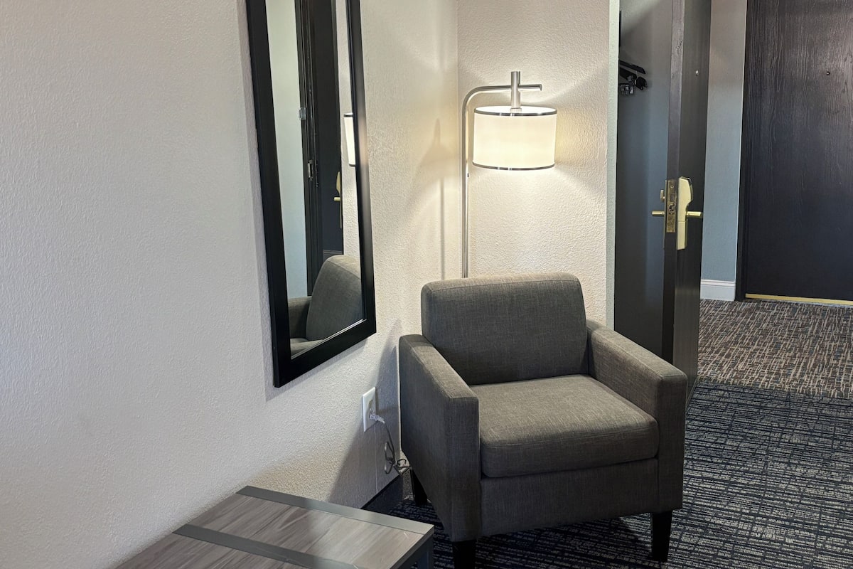 A hotel room with a grey armchair, a standing lamp, a mirror, and a door.