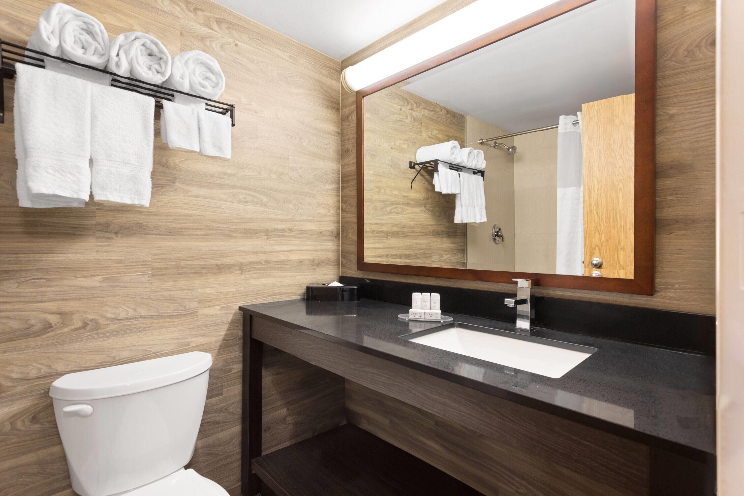 A modern bathroom with a wooden wall, a toilet, a sink with a mirror, and towels on a rack.