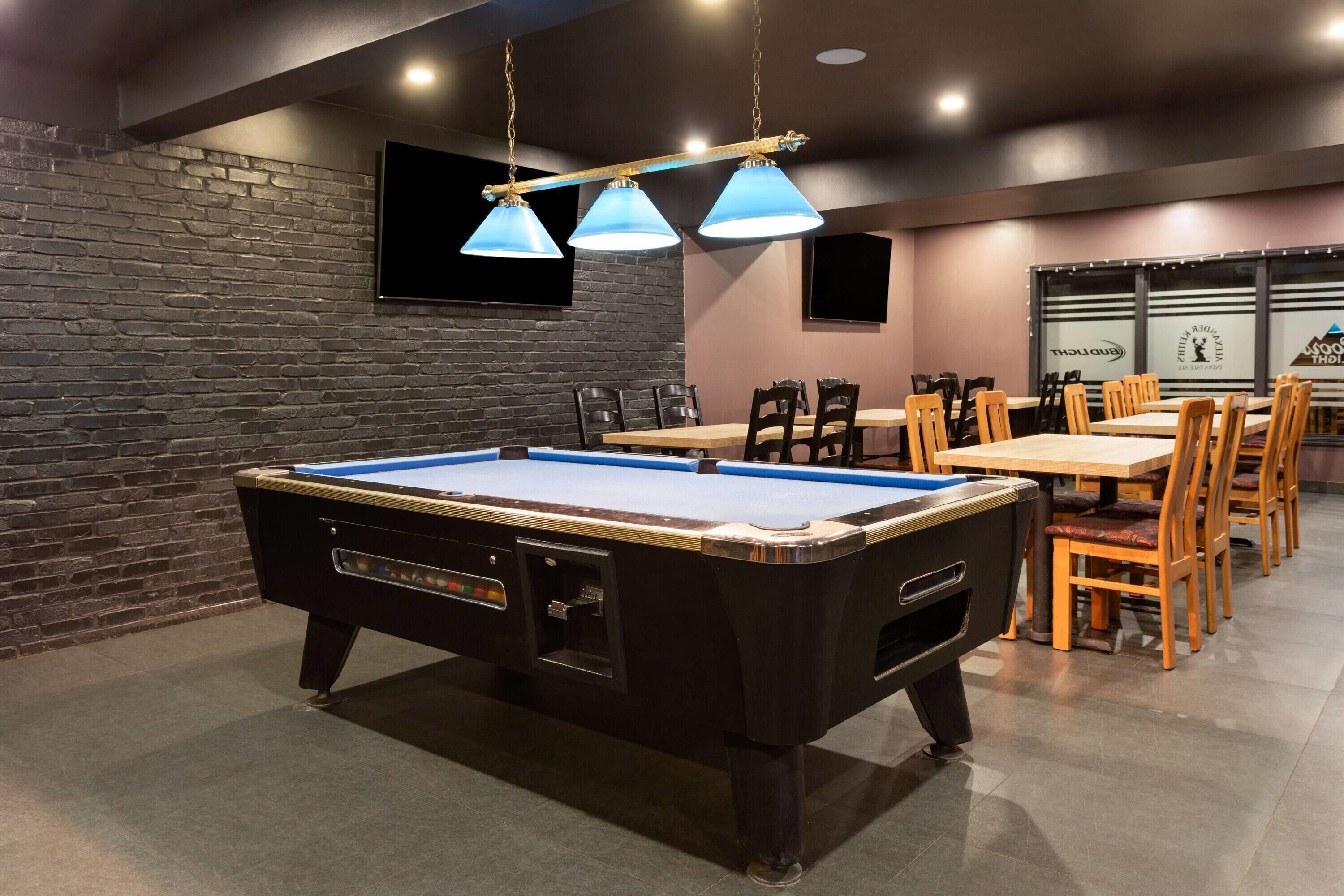Une table de billard éclairée de lumière bleue, un mur en briques et un coin repas avec des chaises et des tables en bois.