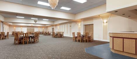 A spacious banquet hall with multiple round tables and chairs, a chandelier, and a reception desk.